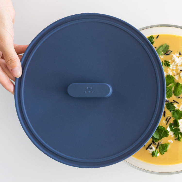 A hand removes a Blue Misen Universal lid from a glass container holding yellow soup topped with parsley, cheese, and pepper.
