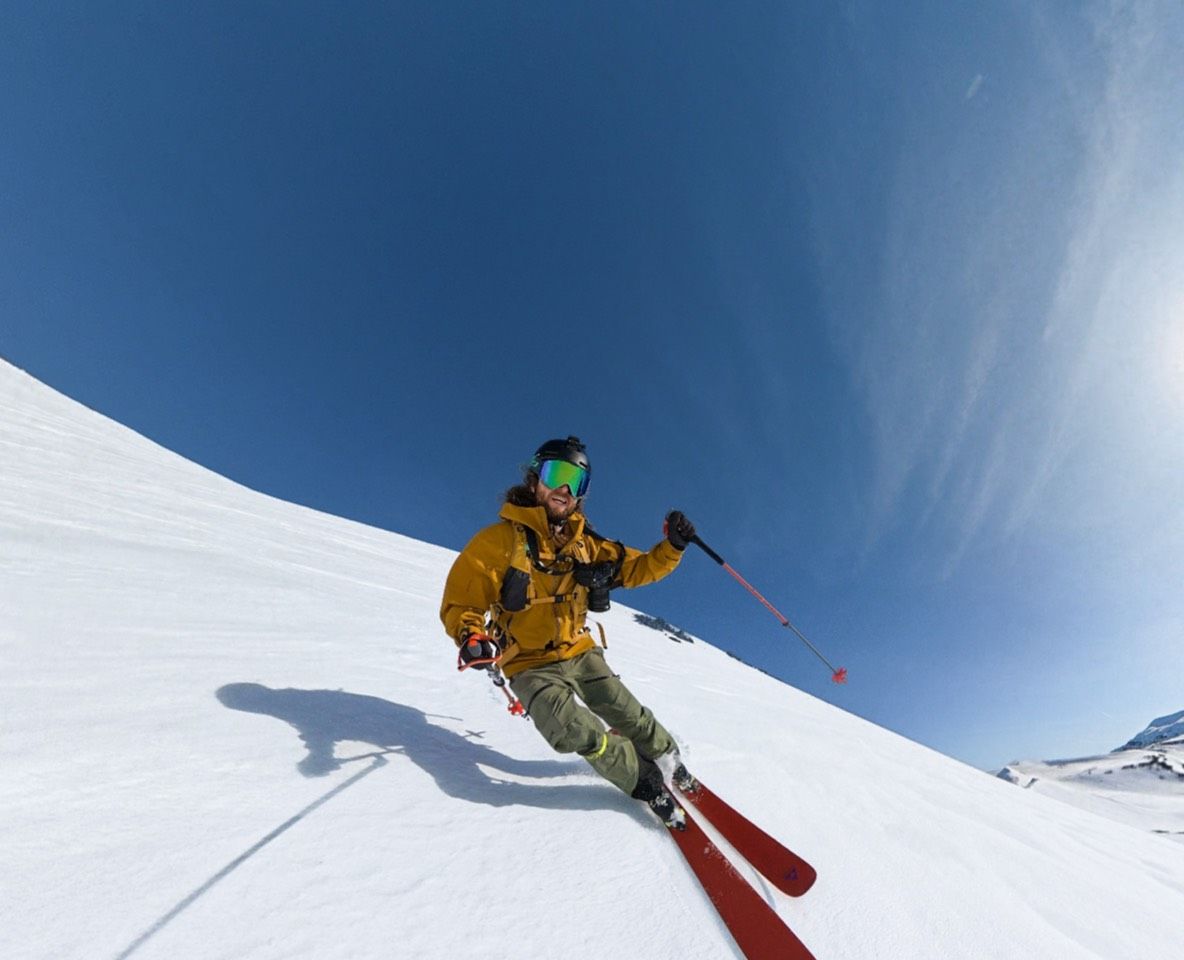 Skier in yellow jacket skiing down a snowy slope under a blue sky, a DJI Osmo 360 II records the descent with an invisible selfie stick