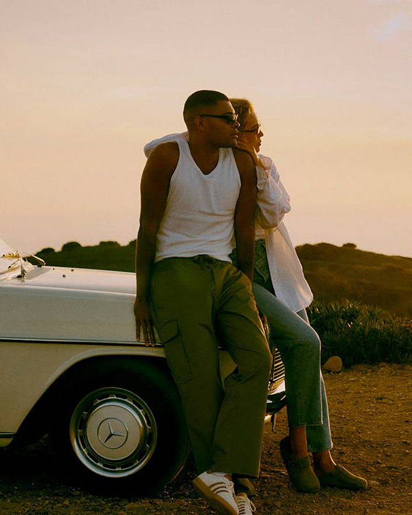 A man and a woman leaning on a car watching the sunset wearing sunglasses.