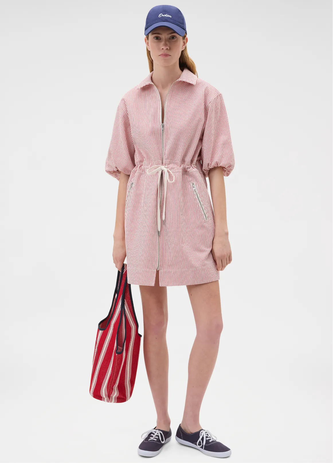 A model in a pink dress with a belt, holding a striped bag.