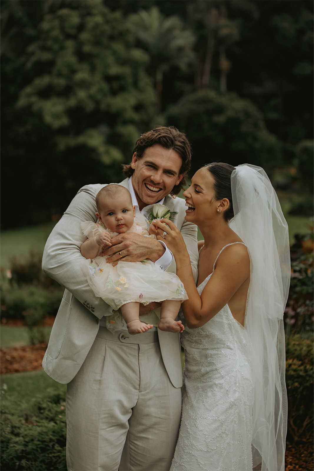A joyful couple holds their baby in a garden setting, celebrating together.