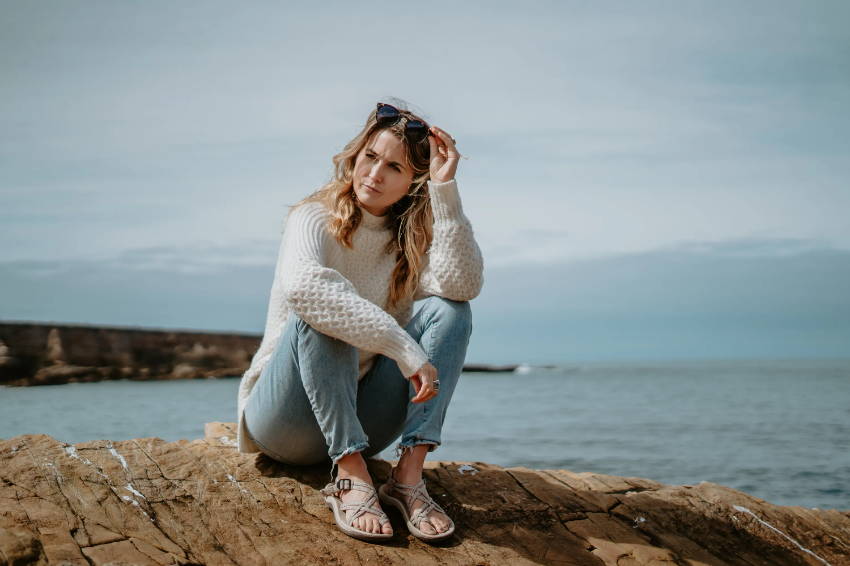 woman sitting on rocks near ocean wearing suppination sandals