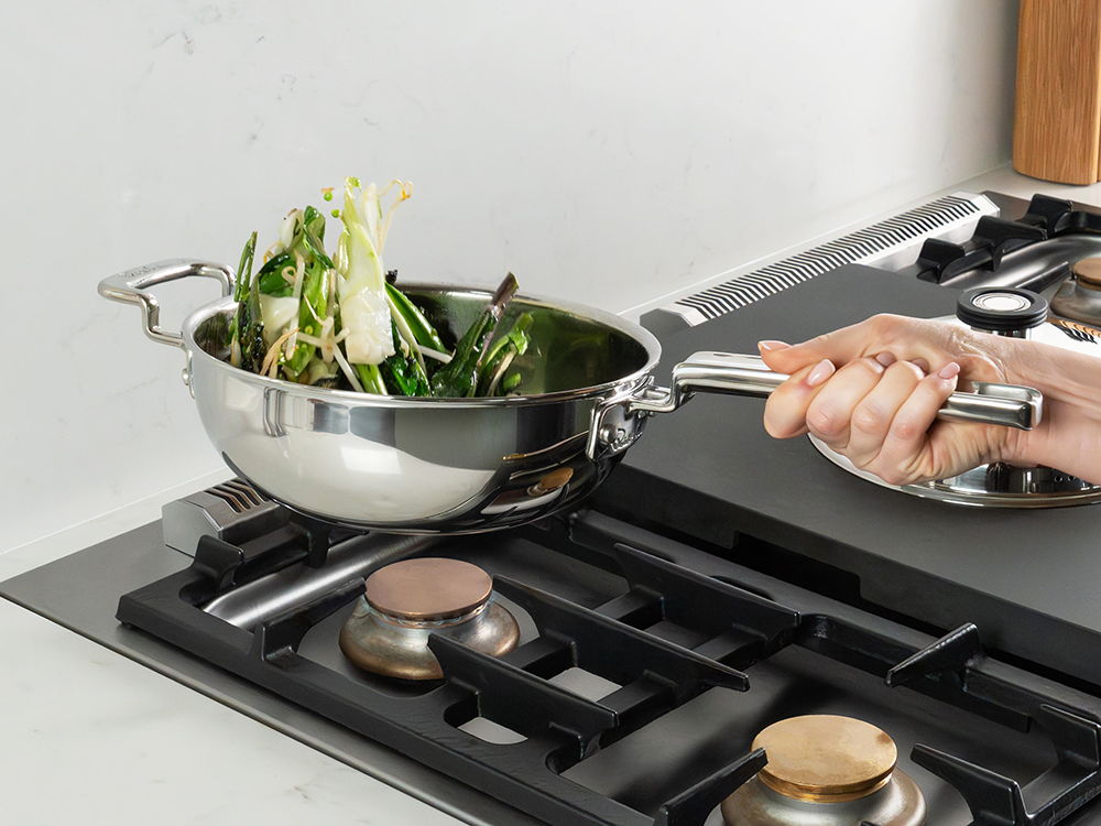 A hand holding 316 Series wok with greens over a stovetop.