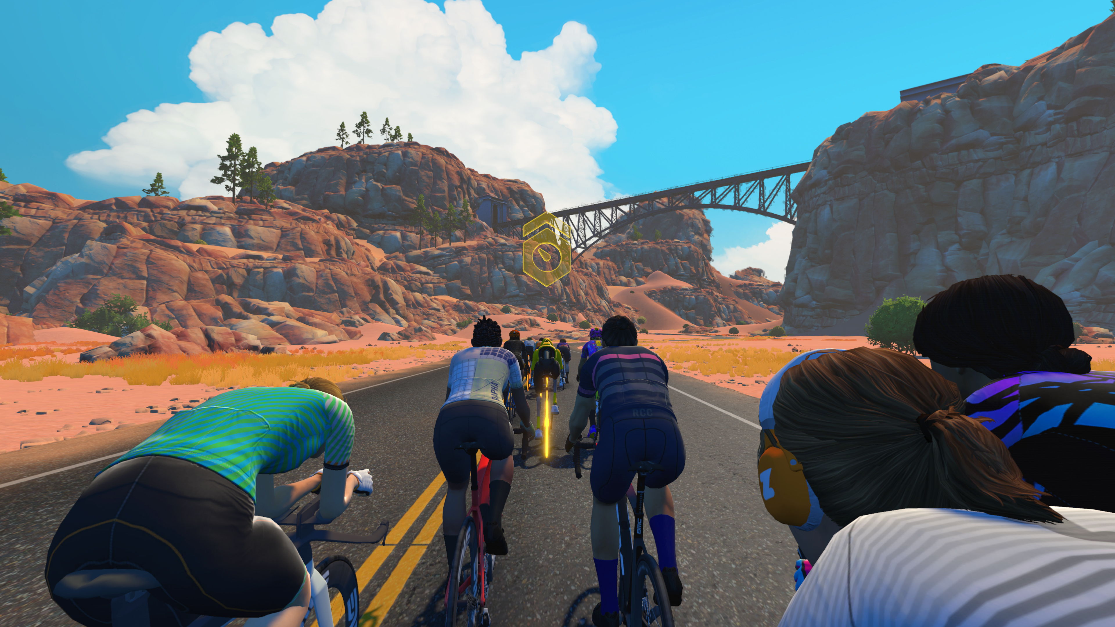 Gruppe von virtuellen Radfahrern fährt in einer Zwift-Umgebung durch eine Wüstenlandschaft mit Felsen und Brücke im Hintergrund.