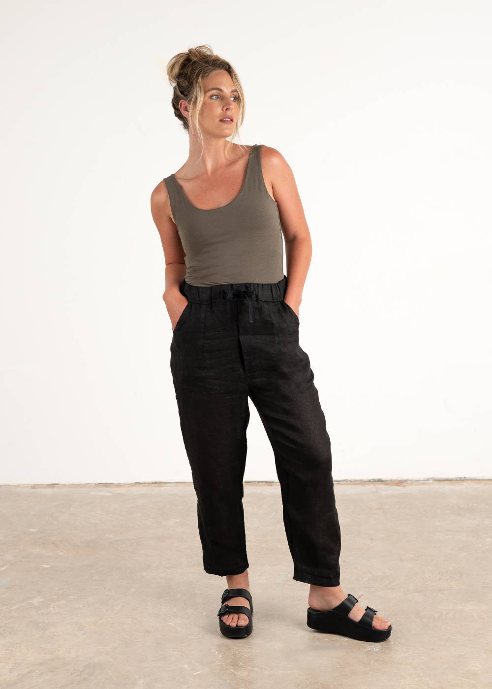 A model wearing a pair of black linen trousers with a khaki green vest and black chunky platform slides