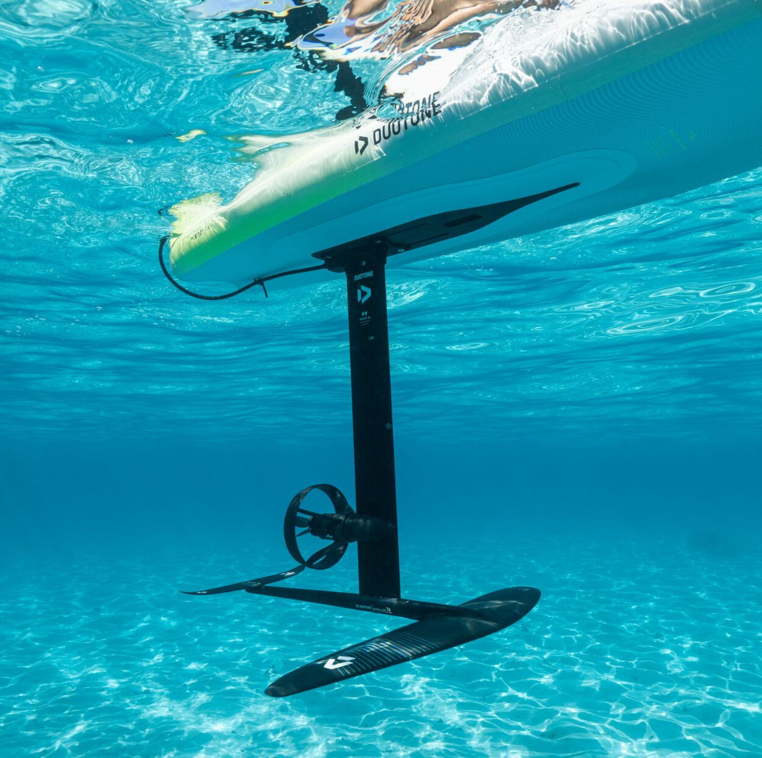 A e-foil submerged underwater beneath a surfboard in clear blue water.