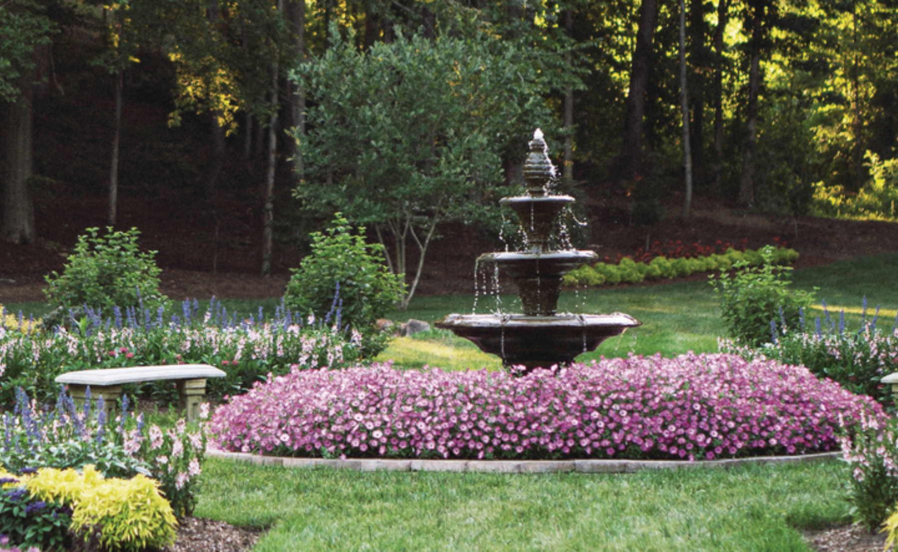 Water fountain surrounded my pink annuals planted in a garden with a bench surronded by more annuals