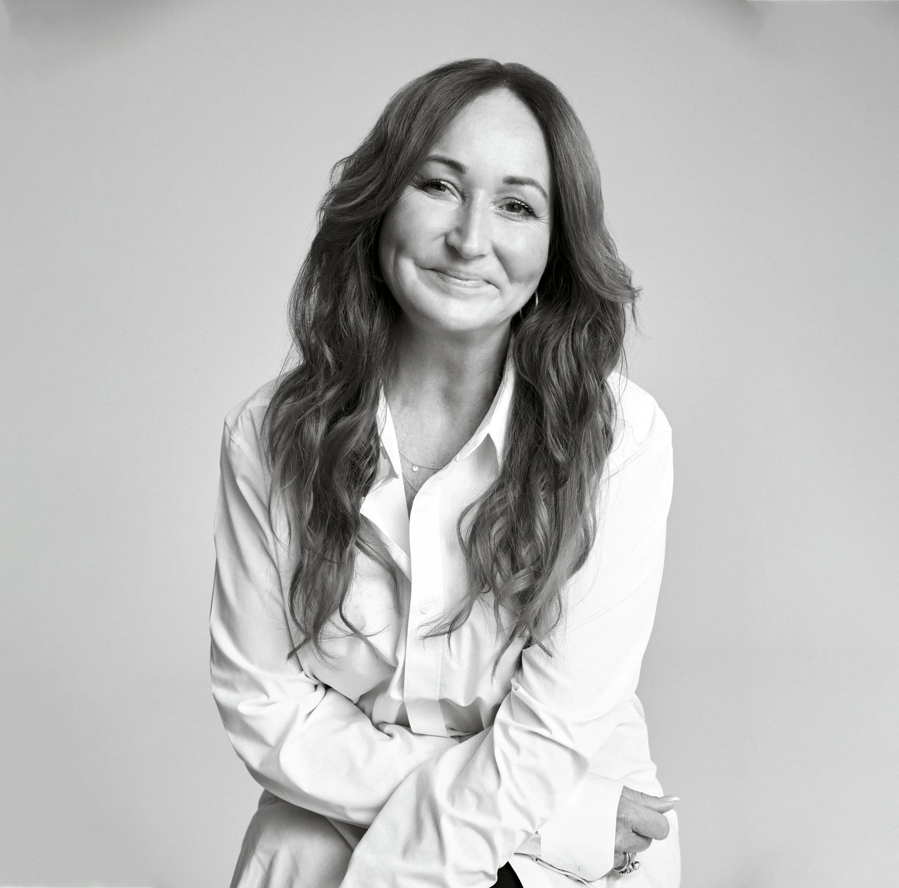 A smiling woman with long hair wearing a white shirt, arms crossed.
