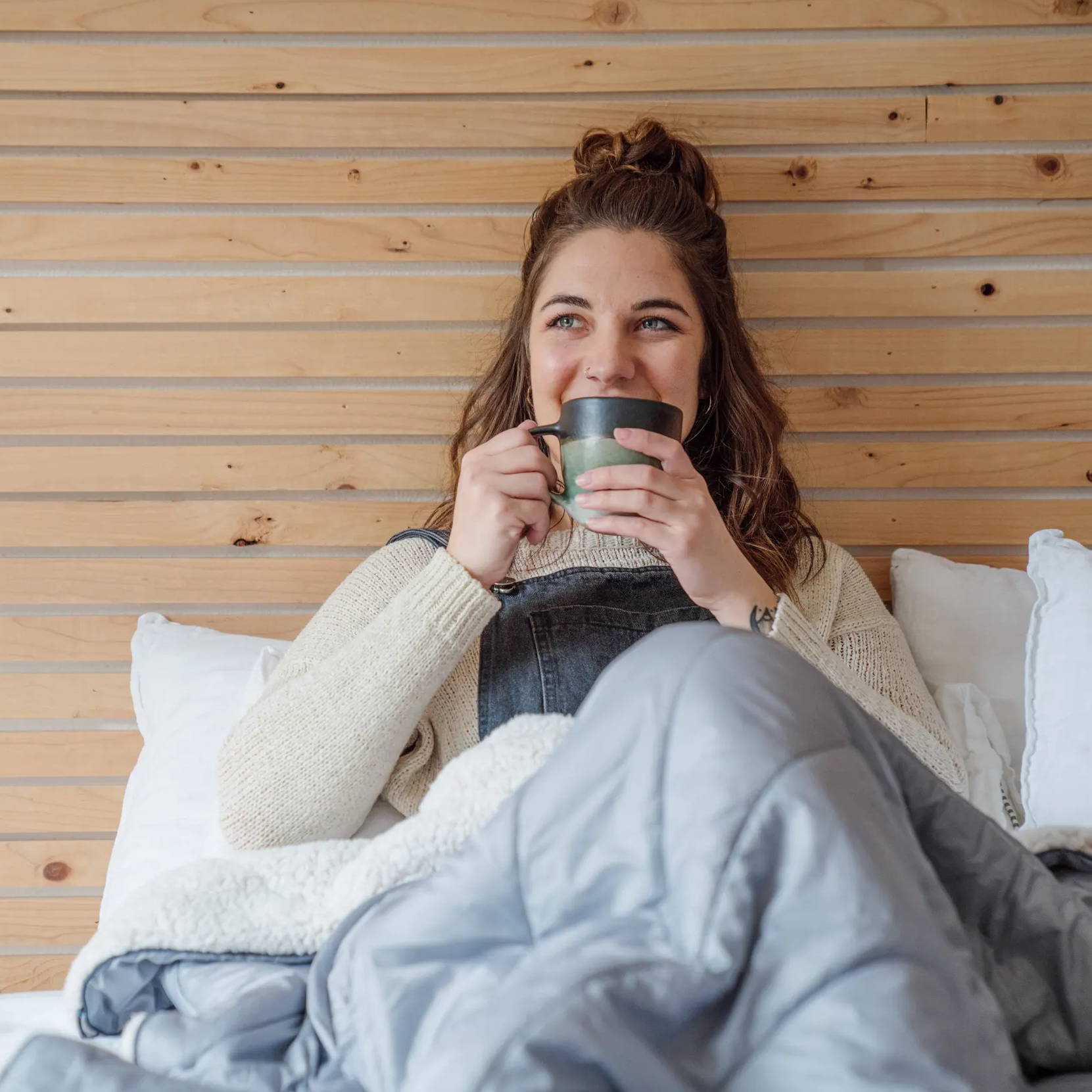 Woman sitting in here bed wrapped in a sherpa fleece blanket drinking coffee