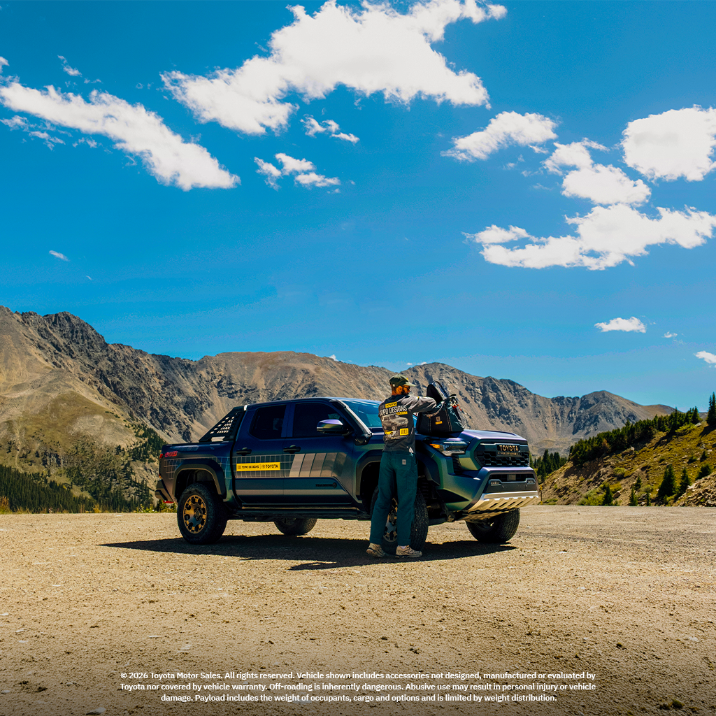 Person standing beside a truck in a mountainous landscape under a blue sky.