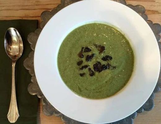 Bowl of green soup with dark garnish and spoon beside it.