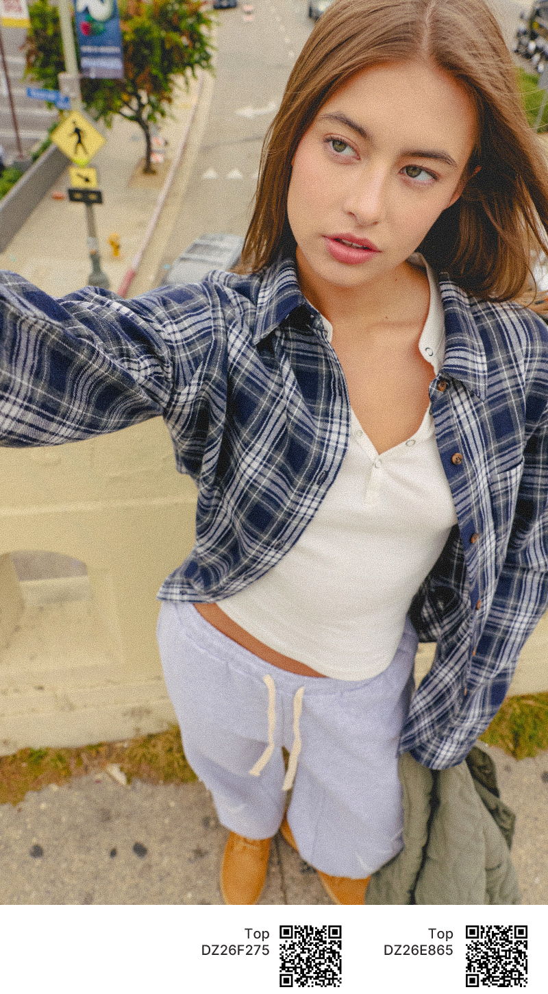 A person taking a selfie outdoors, wearing a plaid shirt and shorts.