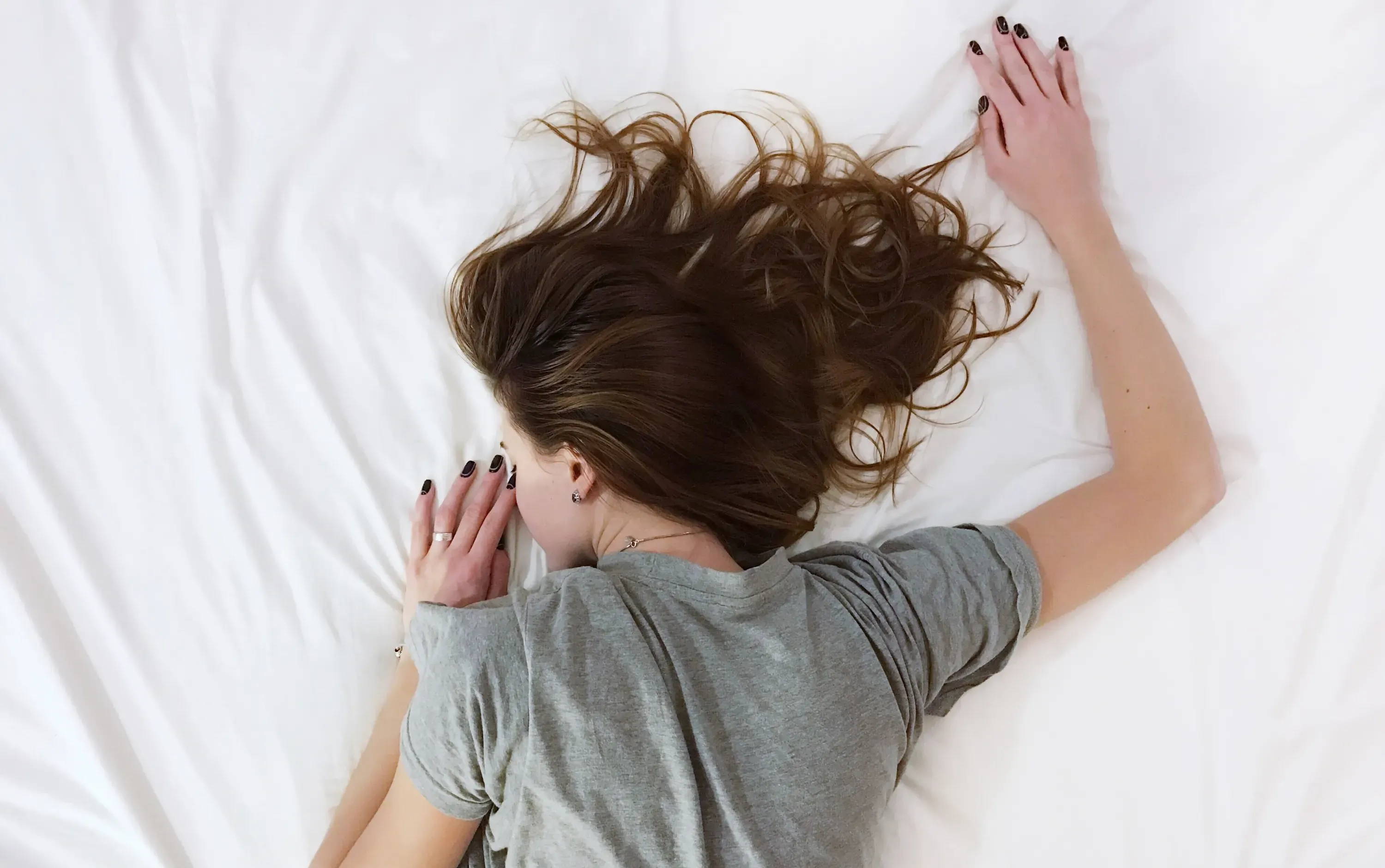 A woman lays face down on a white be in a gray shirt.