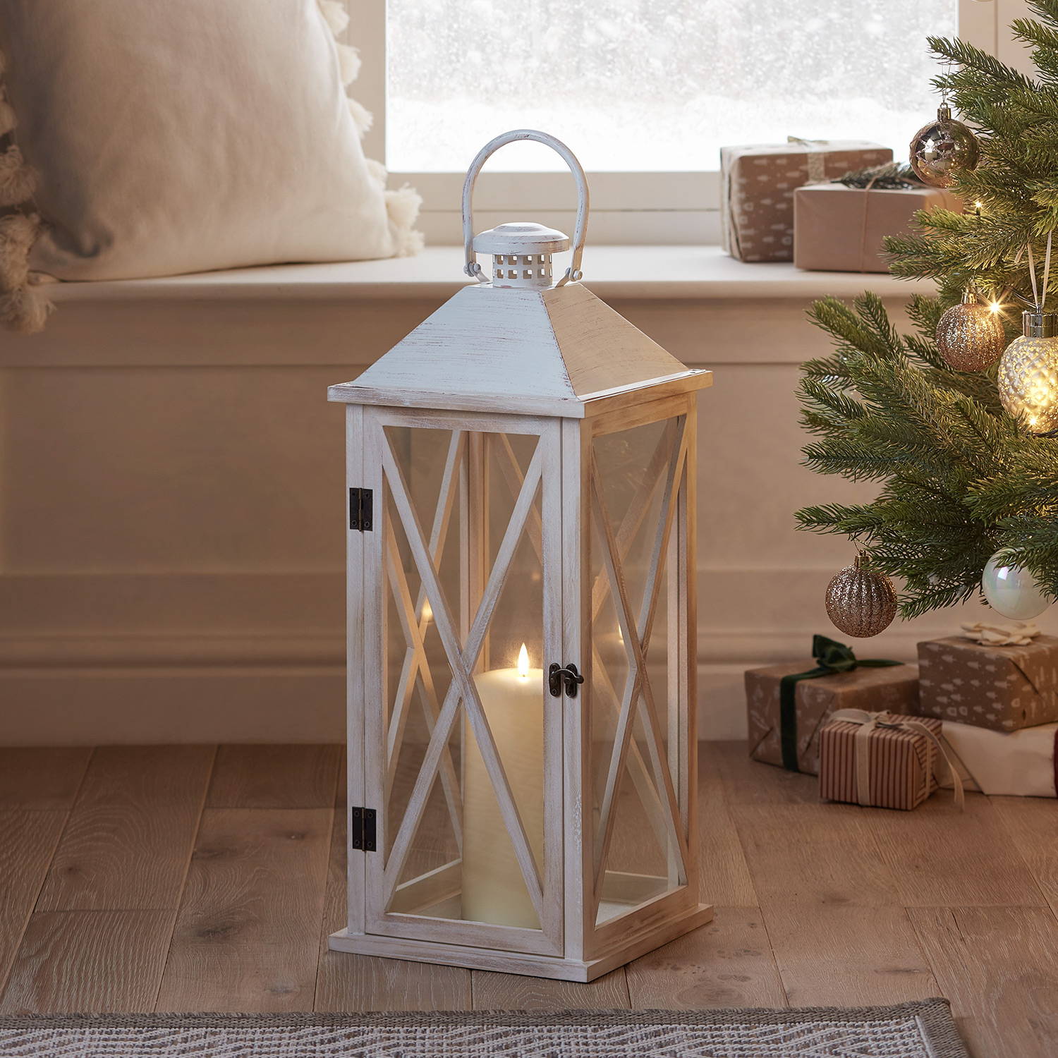 A white wooden lantern with an LED TruGlow candle inside with a cosy Christmas tree in the background.