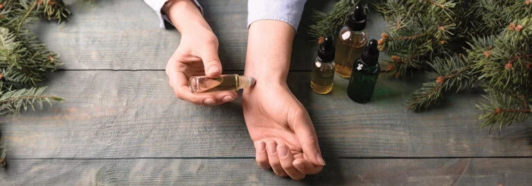 Person applying herbal oil on wrist surrounded by greenery and essential oil bottles.