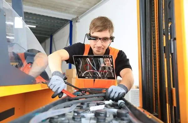 Young man in black T-shirt, orange overalls, and gloves, wearing Vuzix smart glasses while fixing machinery with a tool.