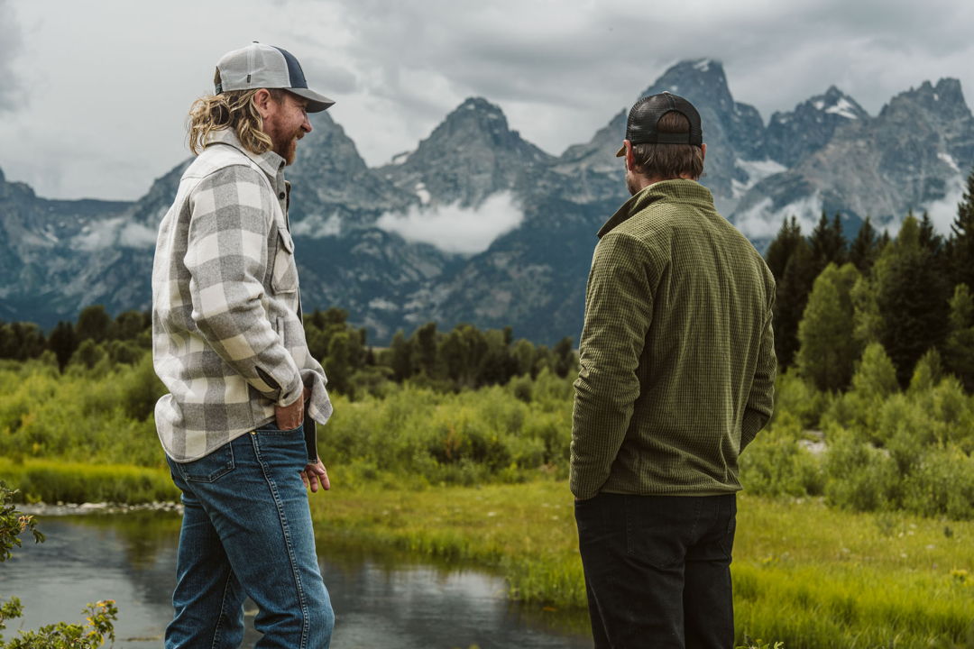 Two men by a mountain river beneath the Tetons, one wearing a Buffalo Jackson plaid flannel shirt jac