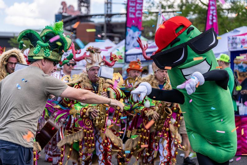 Dilly the Pickle® mascot dancing with a Mummers string band and a fan in a pickle jester hat during the Pickle Parade at The Big Dill® Philadelphia pickle party, with Citizens Bank Park in the background