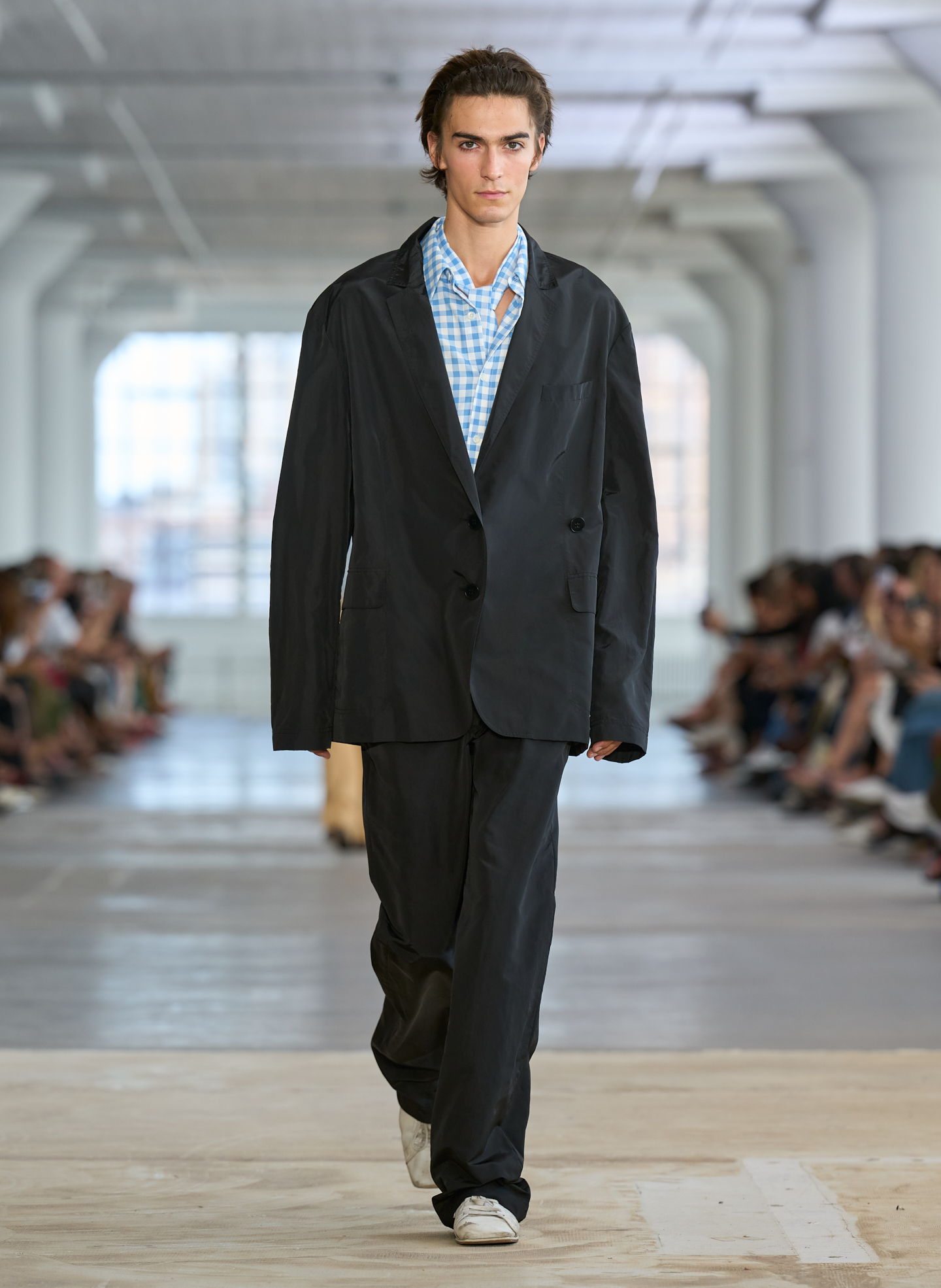 Model walking down runway in oversized black suit and checkered shirt.