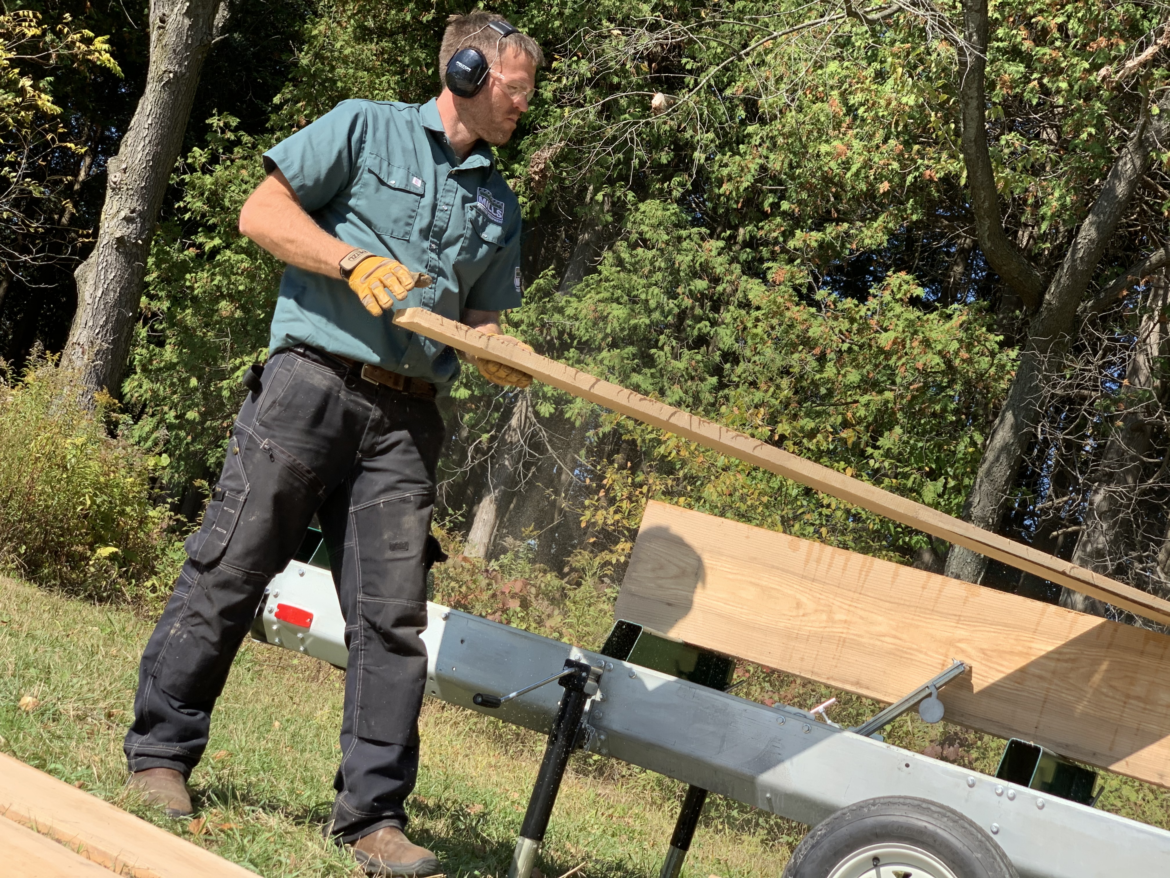 Man wearing headphones, lifting wood plank near trailer in wooded area.