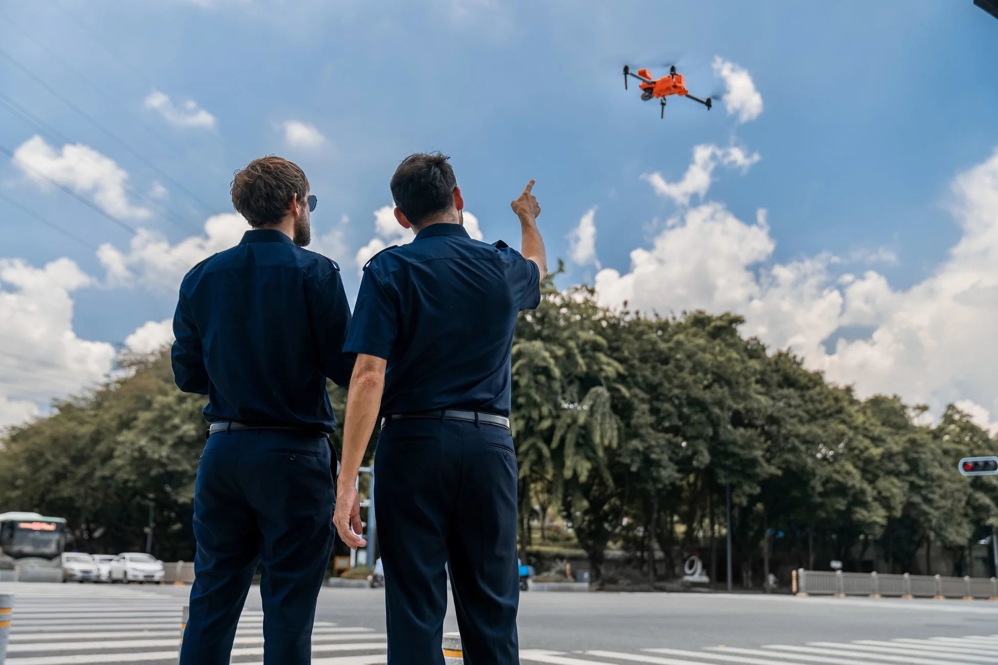 Two men in uniform observing a drone flying in a clear sky.