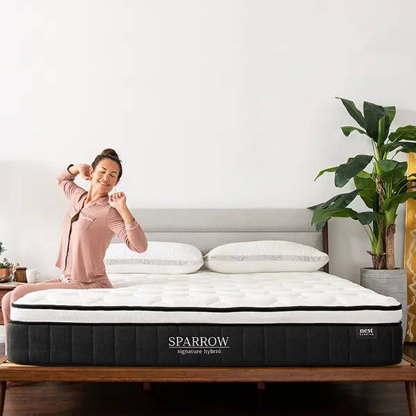 a woman in pajamas sitting and stretching on the side of a sparrow signature hybrid mattress