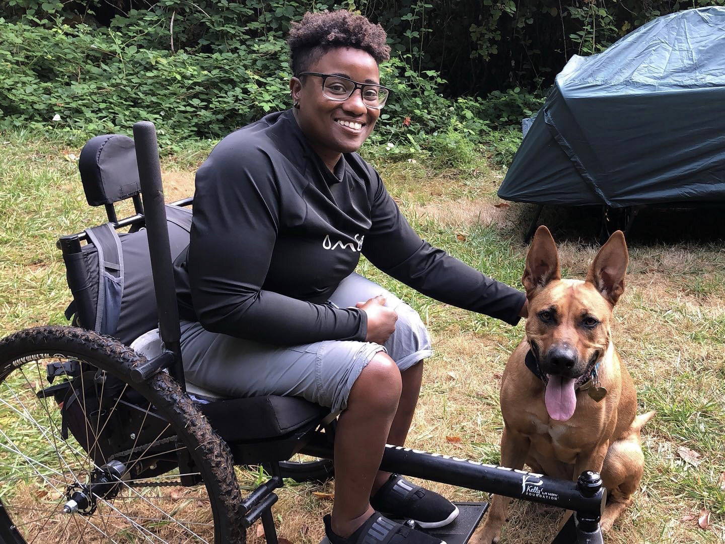 Evita smiling in GRIT Freedom Chair with dog on grassy campground