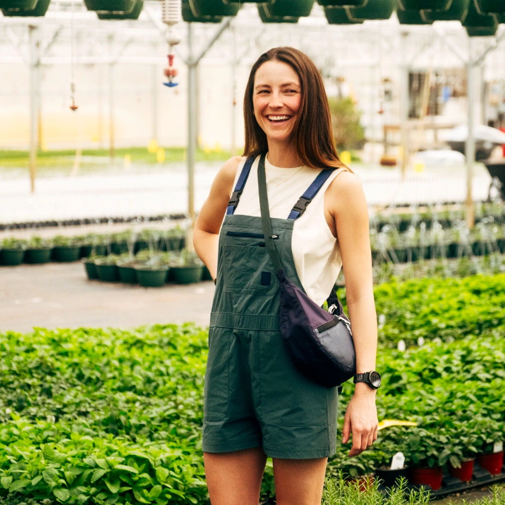 Smiling woman in overalls standing among plants in a greenhouse.