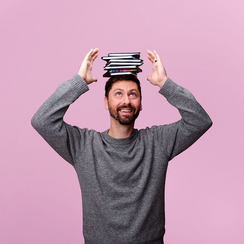 Man smiling with books balanced on his head against a pink background.