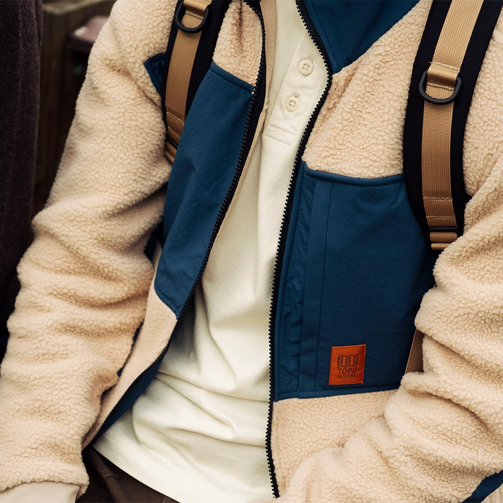 Person wearing a beige and navy jacket, seated with a backpack.