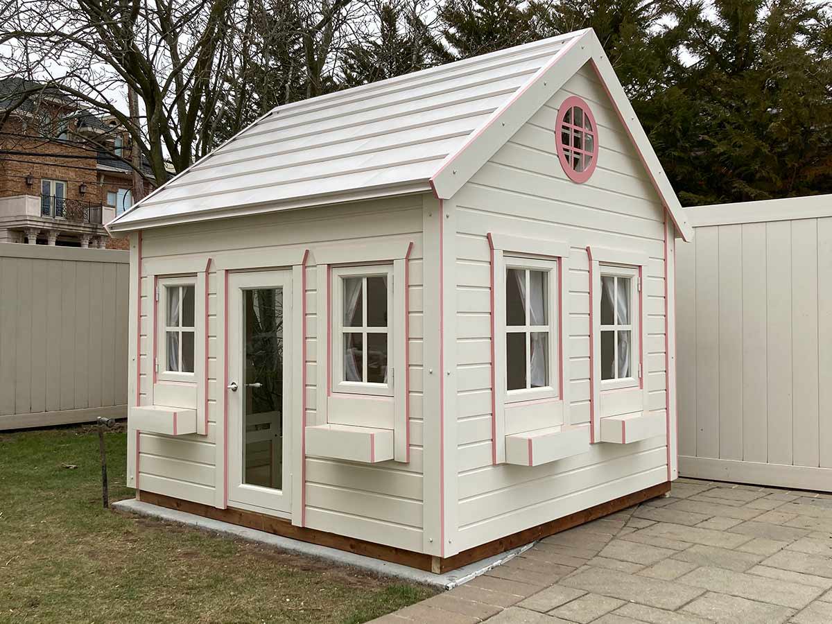 White wooden playhouse with light pink features, glass doors and a round top window in the backyard by WholeWoodPlayhouses