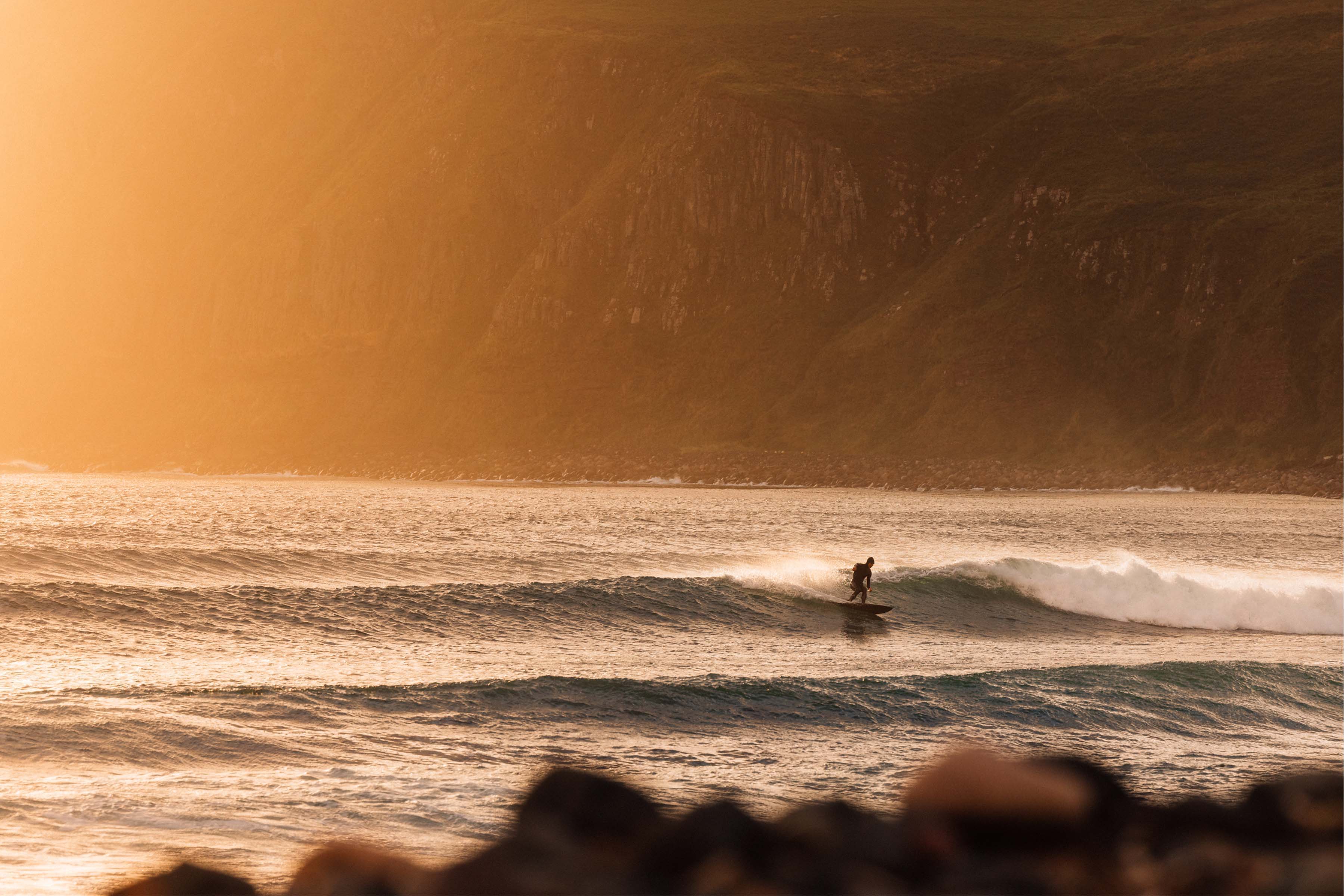 Surfer riding a wave during sunset with rocky shore in the foreground.