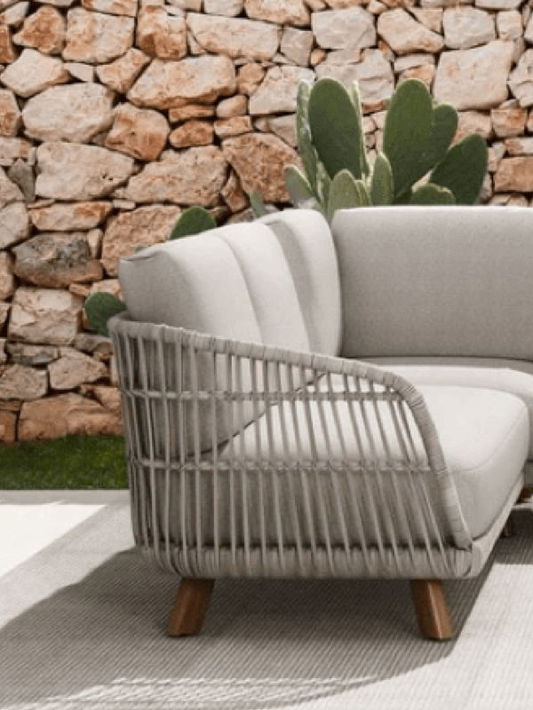 Woven outdoor sectional with matching ottomans on a patio, styled with neutral cushions against a rustic stone garden wall.