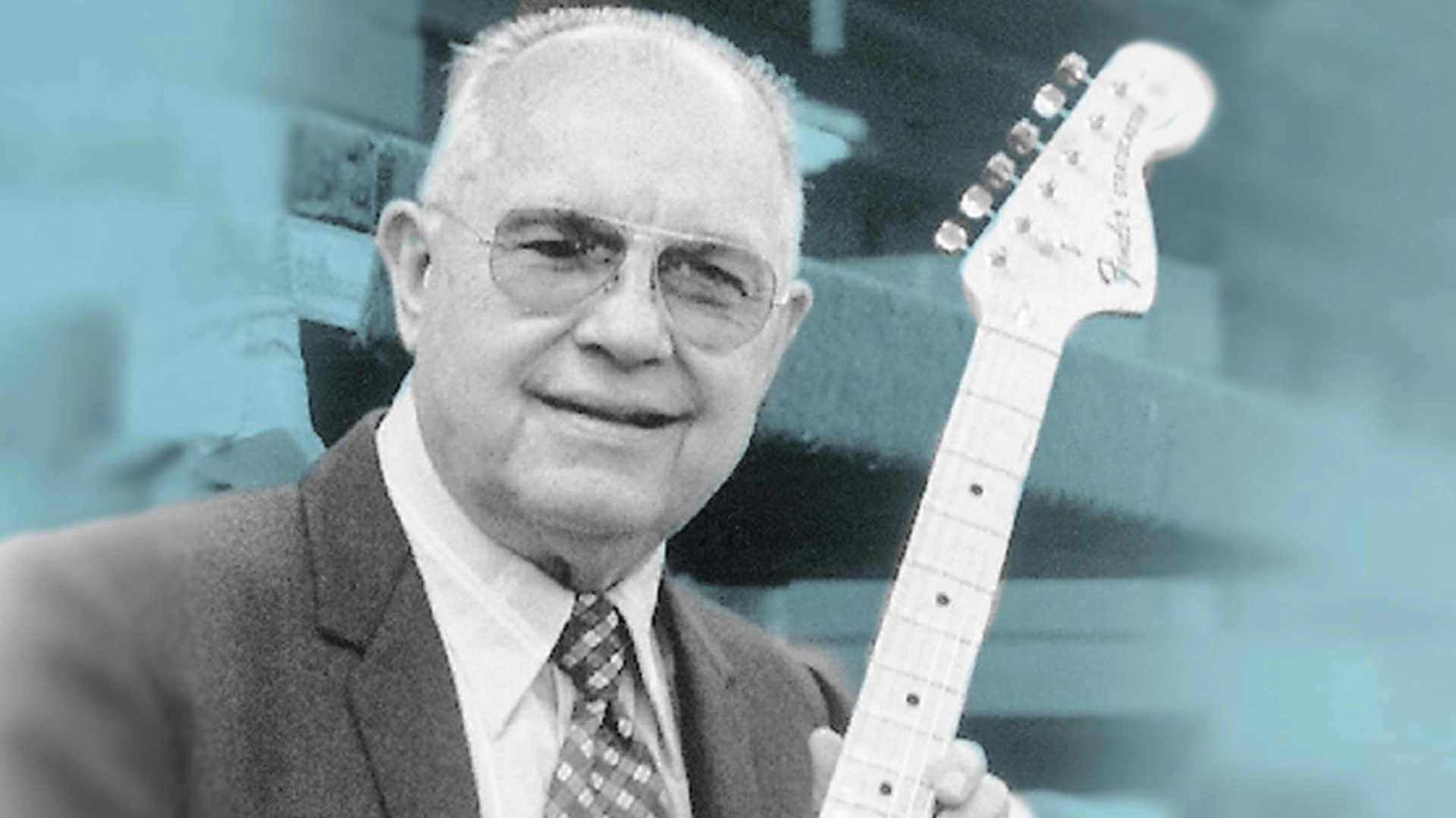 Smiling Leo Fender in a suit holding a Stratocaster electric guitar.