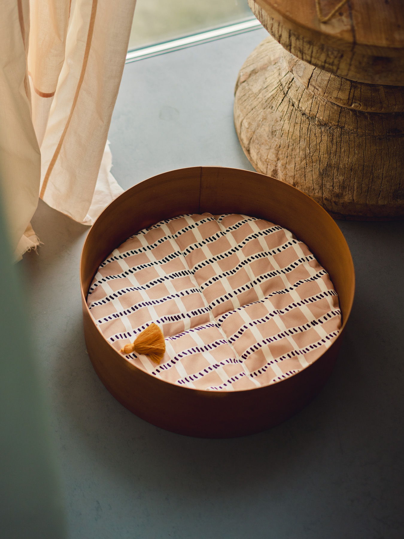 A round pet bed with a patterned cushion near a window.