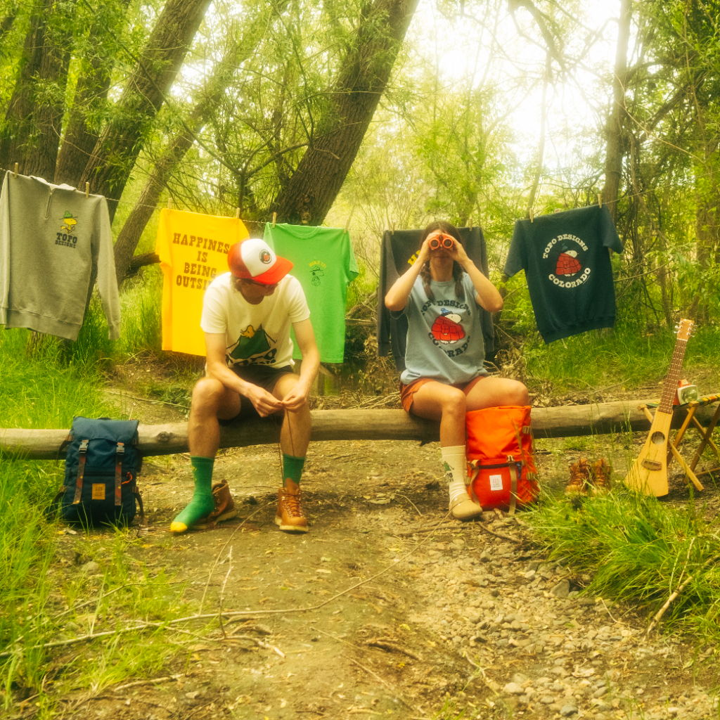Two people sitting on a log with colorful shirts hanging in the background.