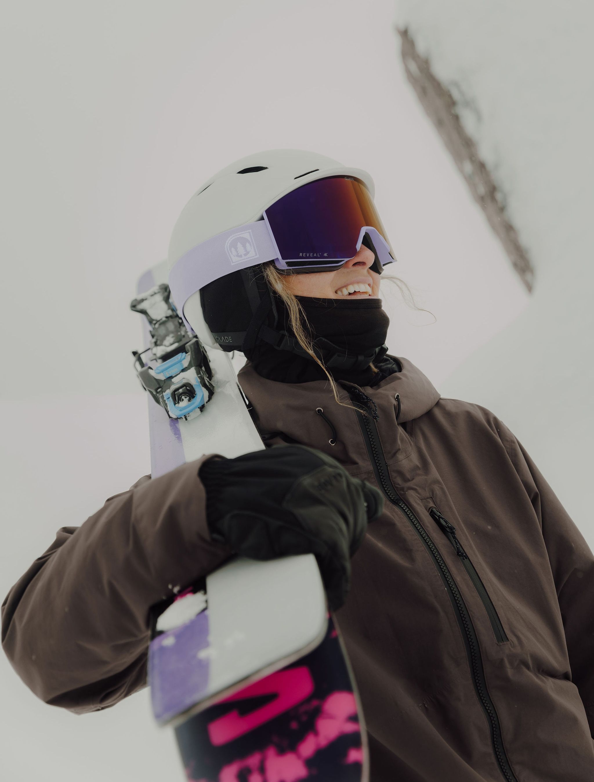 A snowboarder smiling while holding a snowboard in snowy terrain.