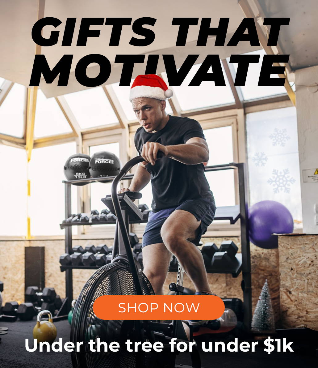 Man exercising on an Air Bike in a home gym, surrounded by gym equipment and festive Christmas decorations, with text overlay: 'Gifts that Motivate - Find the Perfect Fitness Present Under Your Tree for Under $1k.