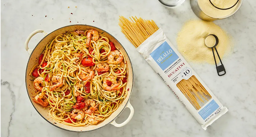 DeLallo Bucatini in package on a marble table next to a pot of prepared pasta with shrimp