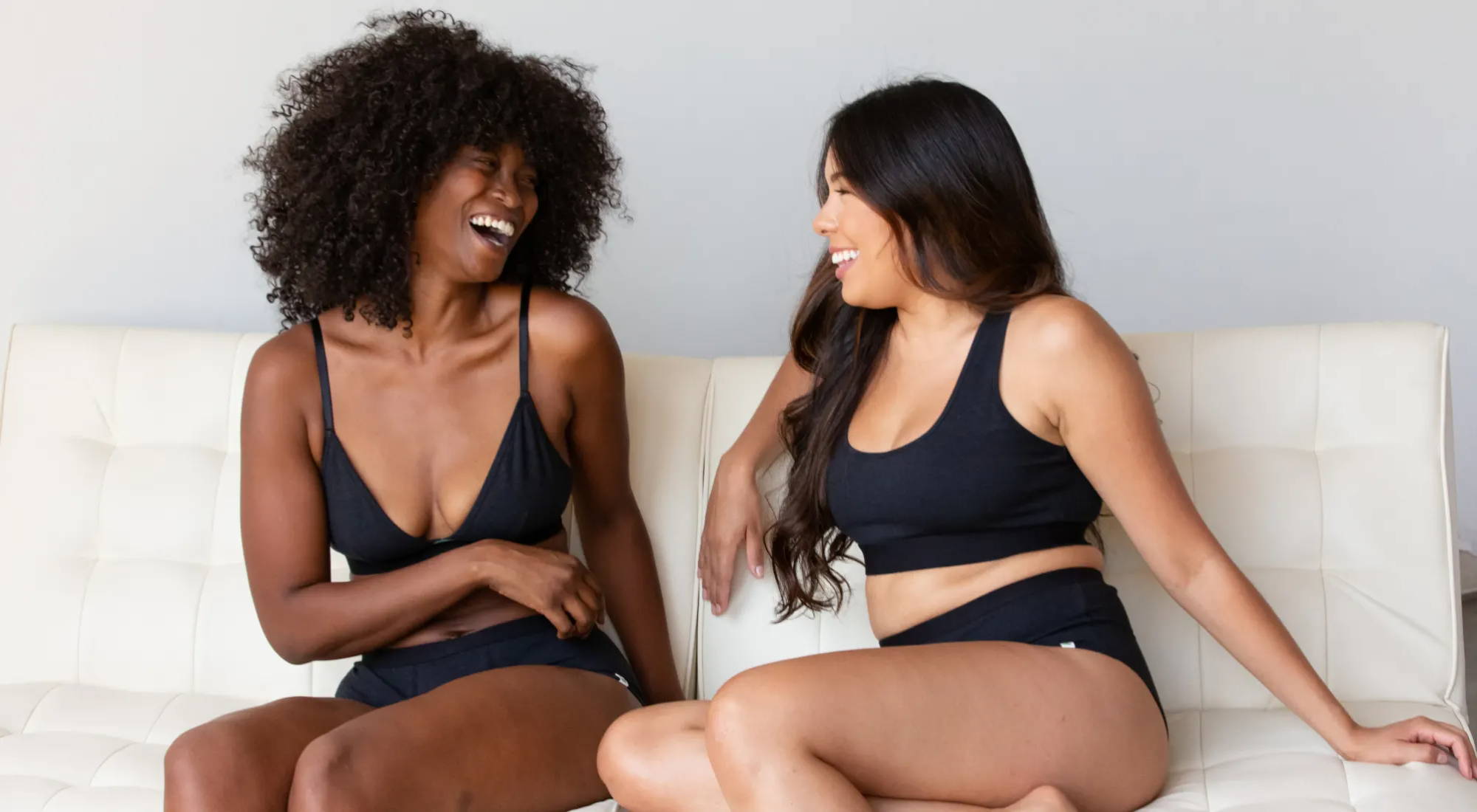 Two women in WAMA hemp bralettes and organic underwear sit on a white couch, laughing.