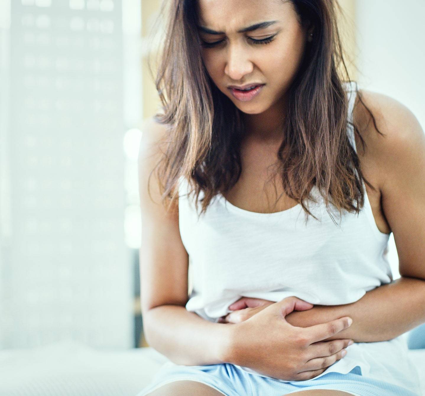Frau mit Symptomen einer Nahrungsmittelallergie, die schmerzverkrümmt auf ihrem Bett sitzt und ihren Magen hält.
