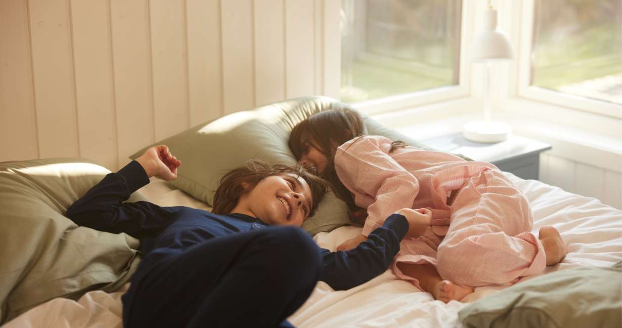 Brother and sister in pyjamas lying on the bed giggling – this is a dust mite hot spot so allergic kids may cough at night