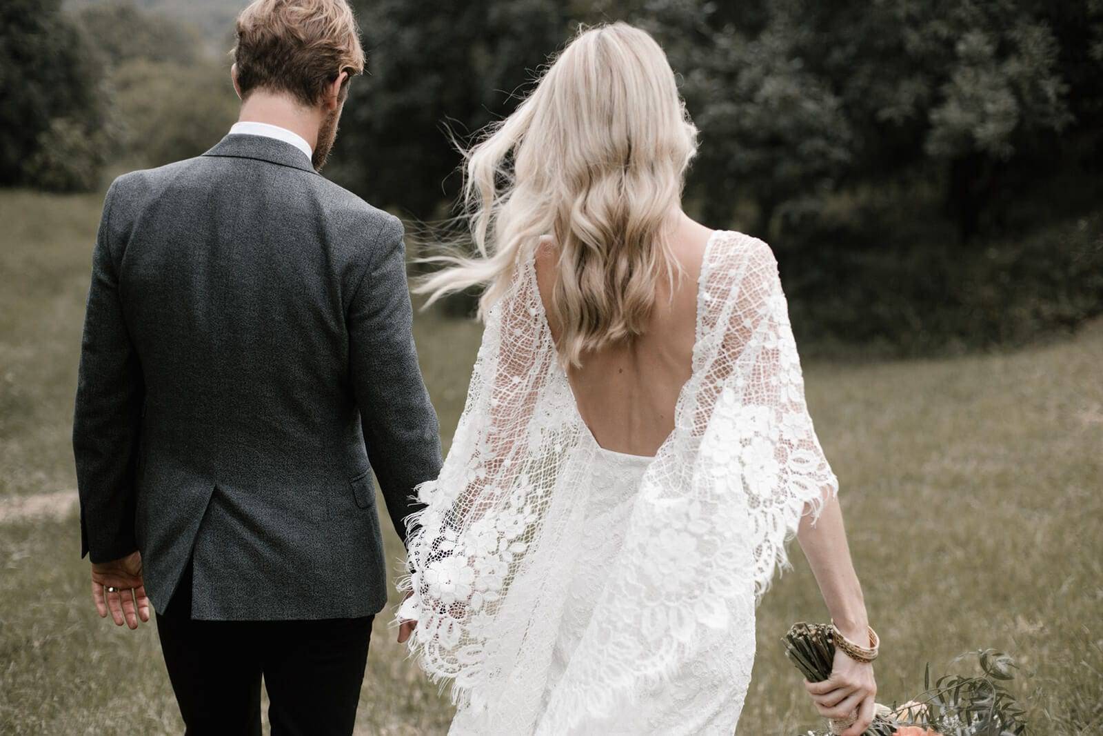 Bride wearing an overthe shoulder lace detail dress walking with her groom 