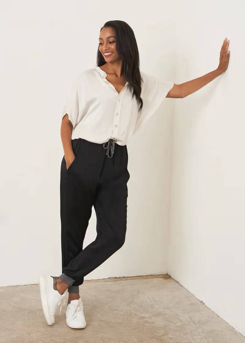A model wearing a white short sleeved shirt with black satin trousers that have glitter detailing on the cuffs and tie waist with white trainers