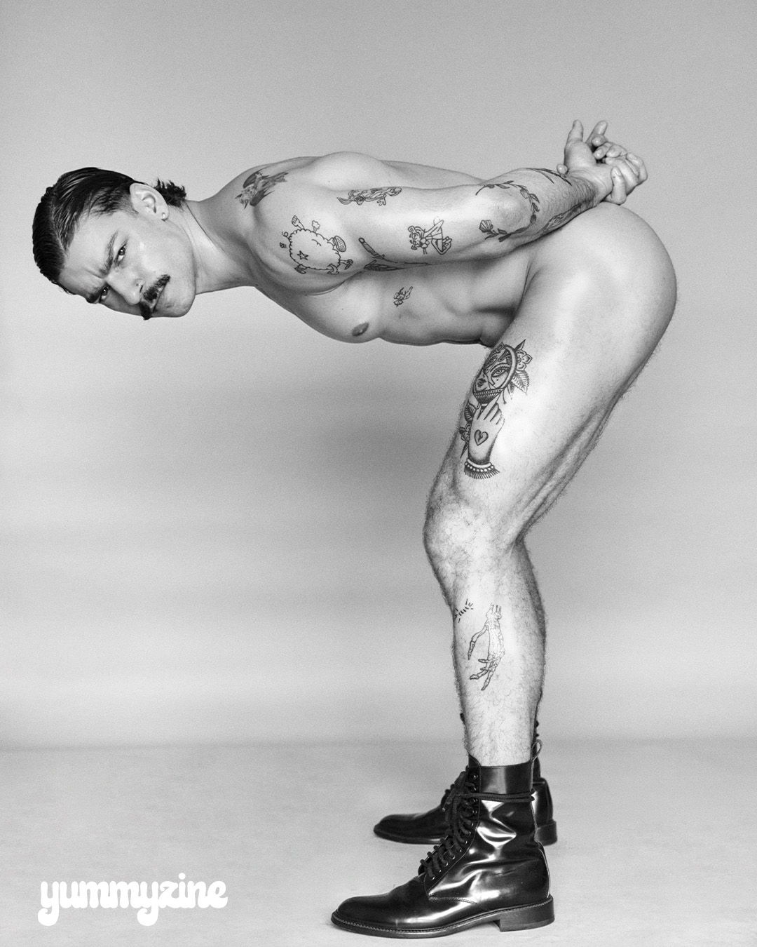 A tattooed man in boots poses with hands behind his back.