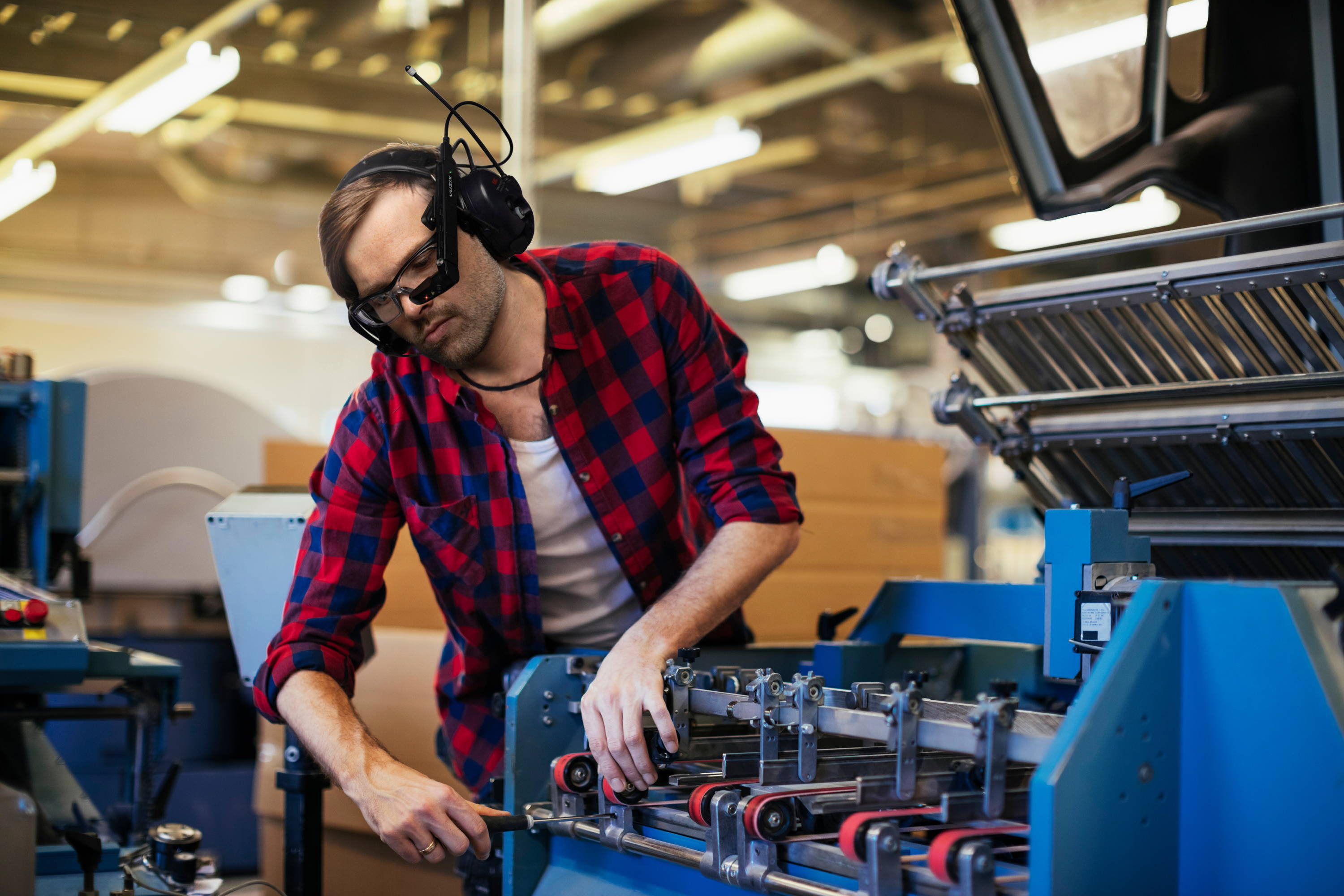 Man in red-and-blue checked shirt, wearing Vuzix smart glasses, repairs machinery on the factory floor.