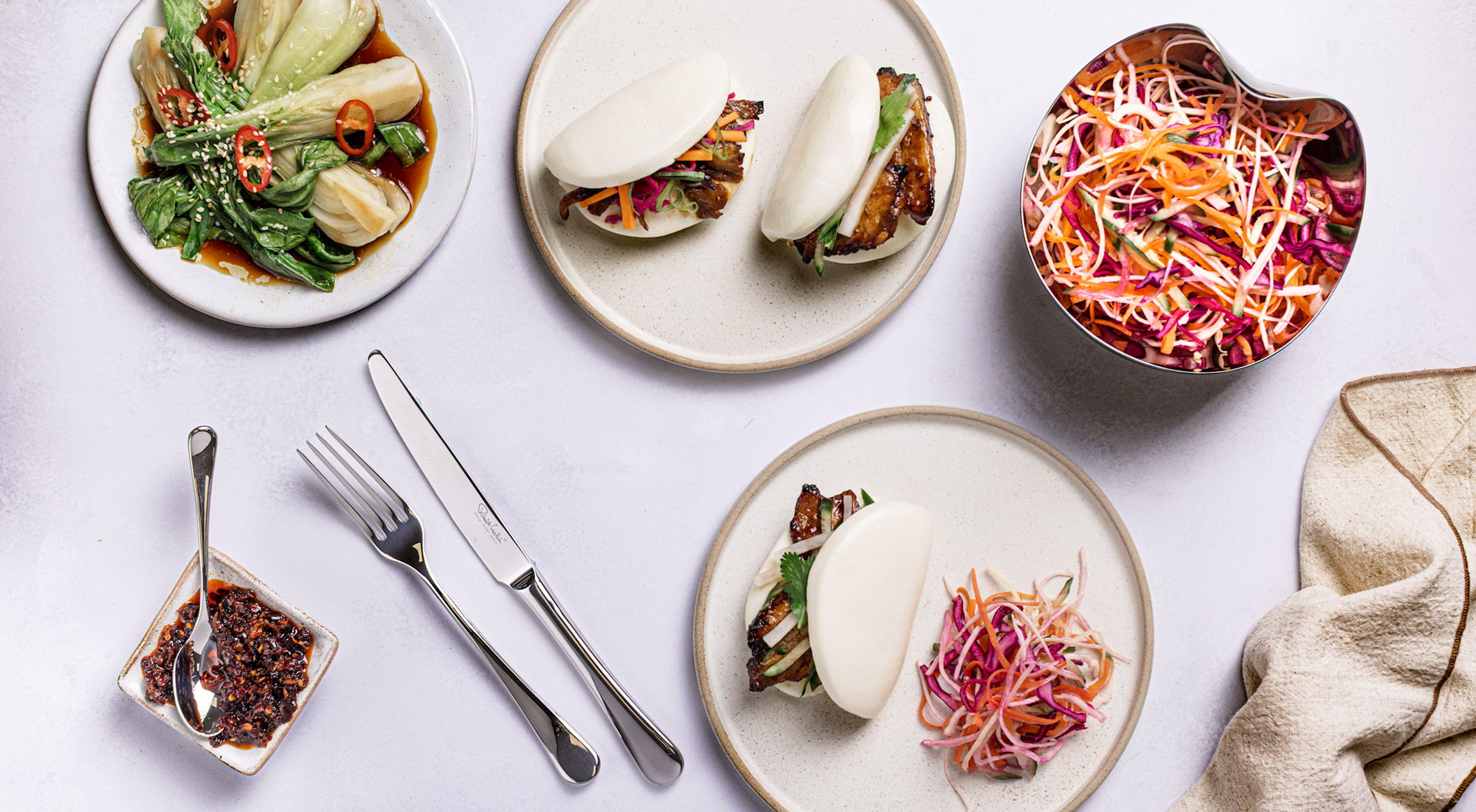 Assorted dishes including pork belly bao buns, salad, and condiments on a table.