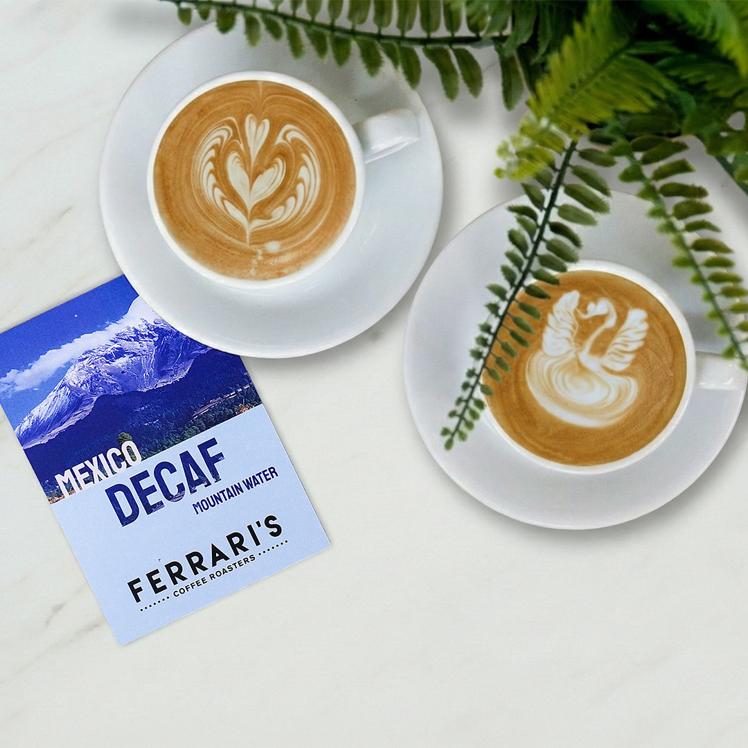 Two cups of latte made with Ferrari’s Mountain Water Decaf coffee beside a Ferrari’s Coffee Roasters decaf card, showcasing smooth crema and rich flavour without caffeine