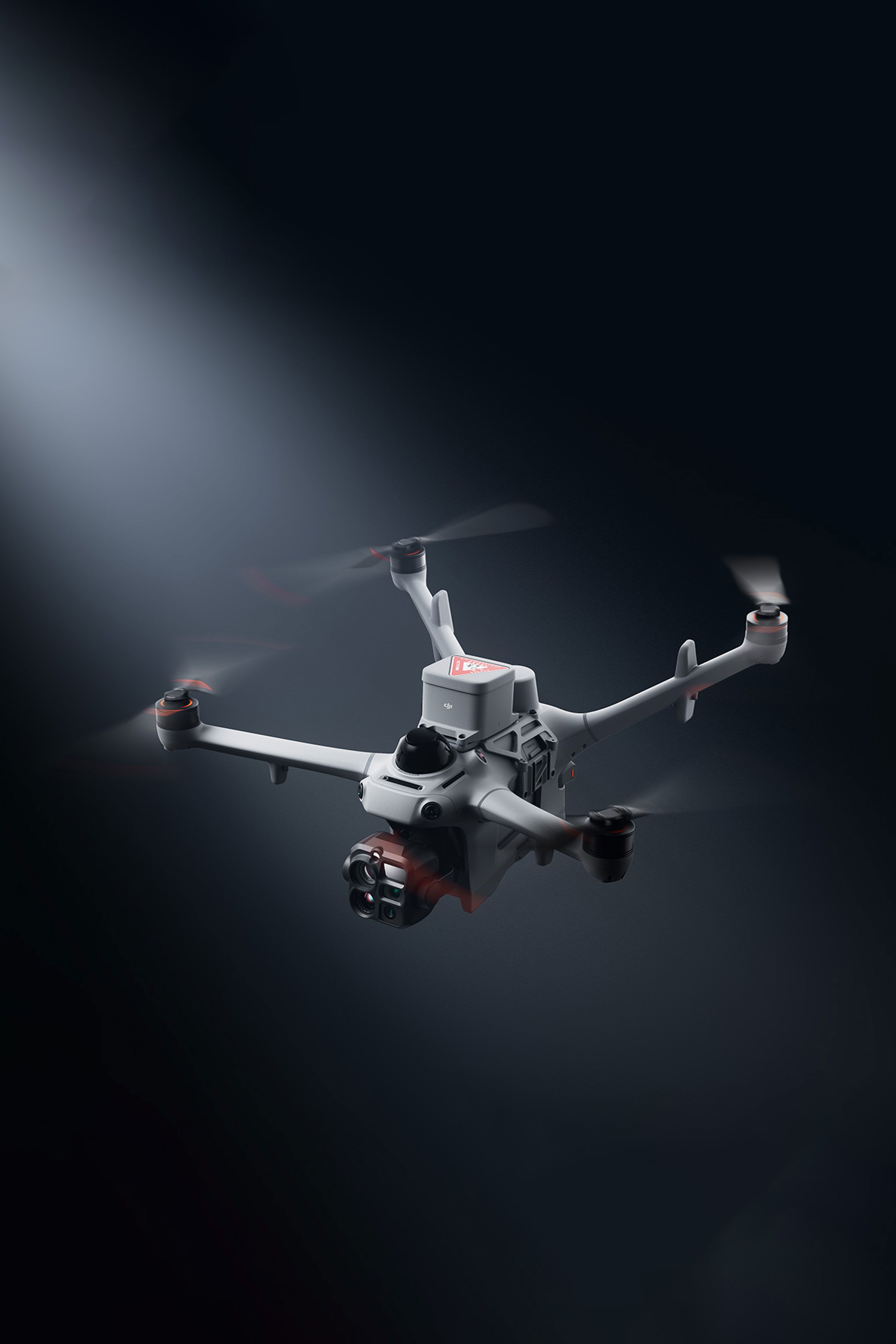 A DJI Matrice 5TD drone flying in a spotlight against a dark background.