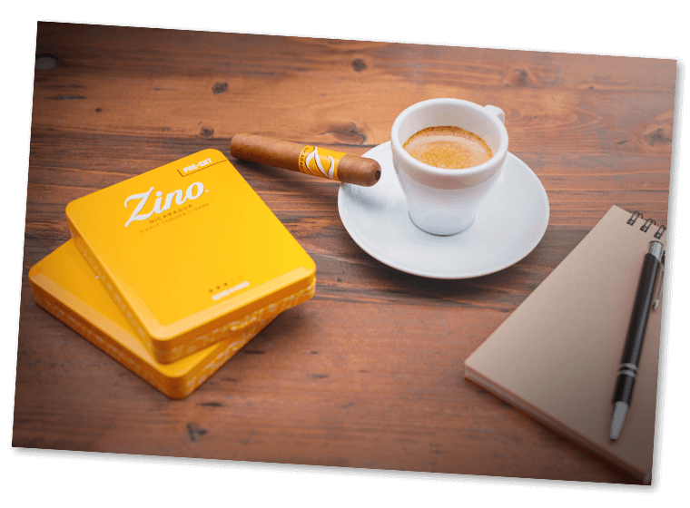 A pre-cut Zino Nicaragua Half Corona resting on the saucer of a filled espresso mug. A notebook, with a pen and two Half Corona tins are placed next to it. 